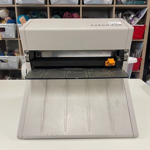 Könnte beinhalten: Ein hellgraues Xyron 850 Klebegerät mit einer breiten Zuführschale, einem schwarzen horizontalen Schlitz und einem kleinen orangefarbenen Kunststoffhebel. Das Gerät verfügt über eine stabile, abgewinkelte Basis und ein rechteckiges Gehäuse.