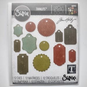 Könnte beinhalten: Ein Sizzix Thinlits Stanzschablonen-Set von Tim Holtz, Modell 662423, mit 12 Metallstanzformen in verschiedenen Etiketten- und Kreisformen. Die Verpackung zeigt die Formen in gedeckten Grün-, Braun- und Rottönen mit dem Text 12 STANZEN.