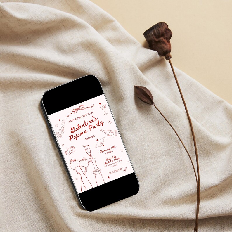 May include: A smartphone shows a Valentine's Pajama Party invitation with red line art illustrations. The invitation includes text, a bow, champagne glasses, and the date February 14th. The phone rests on a beige fabric with a dried flower.
