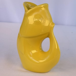 May include: A bright yellow ceramic pitcher shaped like a fish. The vessel features a wide, open mouth at the top serving as the pour spout, a molded eye, and a curved handle formed by the fish's tail. The surface has a smooth, glossy finish.