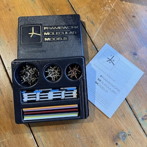May include: A black plastic box set of Framework Molecular Models with a matching instruction manual. The kit contains three circular compartments filled with metal connectors and two rectangular sections holding various colored plastic rods.
