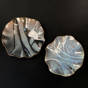 May include: Two ceramic plates with irregular, wavy edges and a glossy finish. The surfaces feature a mottled blend of blue, gray, black, and white tones with a metallic gold-colored trim along the scalloped rims, creating a textured appearance.