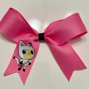 May include: A vibrant pink hair bow made from grosgrain ribbon. The bow has a black centre and features a cartoon cat graphic on one side. The cat illustration is white, yellow, and black.