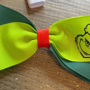 May include: A handmade hair bow featuring a green and lime green layered design. The lime green layer has a black graphic of a character's face. The bow is secured with a red band.