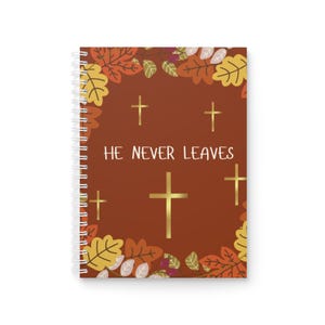 Peut inclure: Cahier à spirales avec une couverture marron ornée de croix dorées et du texte "HE NEVER LEAVES". La couverture est décorée de feuilles d'automne aux tons orange, jaune et marron.