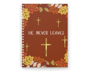 May include: Spiral-bound notebook with a brown cover featuring gold crosses and the text "HE NEVER LEAVES." The cover is decorated with autumn leaves in shades of orange, yellow, and brown.