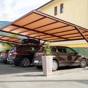 Può includere: Un posto auto marrone con una struttura del tetto rettangolare, che ripara diverse auto parcheggiate. Il tetto è sostenuto da pali di legno. Lo sfondo presenta un edificio e una catena montuosa.