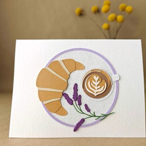 May include: A white card with a paper cutout design of a croissant, coffee cup with latte art, and lavender sprig, within a purple circle. Yellow flower stems are in the background.