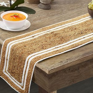 Può includere: Runner da tavolo rettangolare in iuta color beige naturale, con due strisce bianche. Il runner è appoggiato su un tavolo di legno. Sono visibili anche una ciotola bianca di zuppa e una ciotola di legno di pasta.