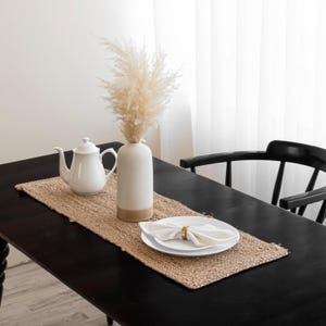 May include: A dining table setting featuring a woven jute table runner. A white teapot and a tall white vase with dried pampas grass are on the runner. White plates with a napkin and gold ring are set on the table. A black chair is in the background.