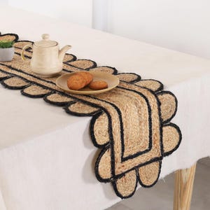 May include: A jute table runner with a scalloped edge and black trim. A beige teapot and plate with biscuits sit on the runner, which is placed on a white tablecloth. The runner adds a rustic touch to the table setting, ideal for a dining room.