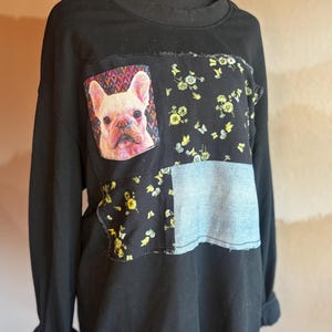 Puede incluir: Sudadera negra con diseño patchwork. Los parches incluyen una imagen de bulldog francés, tela floral con flores amarillas y tela azul claro similar al denim. Las mangas están dobladas.