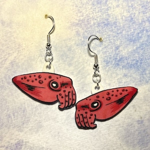 May include: A pair of red and white dangle earrings featuring a cartoon cuttlefish design. The earrings are made of wood and have silver hooks.