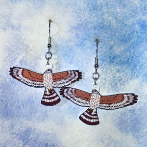 May include: A pair of silver earrings featuring a red-tailed hawk design. The hawk is depicted in flight with brown, white, and red markings.