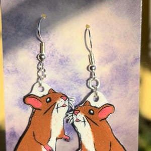 Puede incluir: Un par de pendientes colgantes con una ilustración de dibujos animados de dos ratones marrones y blancos con colas rosas. Los pendientes están hechos de madera pintada y tienen ganchos plateados.