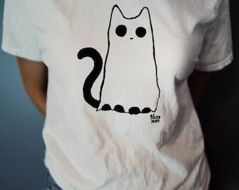 May include: White t-shirt with a black and white illustration of a cat. The cat has large eyes and a long tail. The text "Nerd Paws" is printed below the cat.