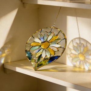 Op de afbeelding: Een set van drie keramische mokken met een gespikkelde, crèmekleurige glazuur. Elke mok heeft een handgeschilderd bloemmotief in blauw, groen en geel. De mokken hebben een eenvoudige, afgeronde vorm.