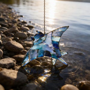 May include: A stained glass manta ray suncatcher in shades of blue, turquoise, and white hangs from a chain. The suncatcher is made of various geometric shapes and is suspended above a rocky shoreline with water in the background.