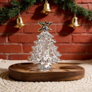 Op de afbeelding: Een heldere glazen kerstboom met gouden accenten, bekroond met een gouden en zilveren ster. De boom staat op een houten dienblad, met gouden bellen en groenblijvende takken op de achtergrond. Een feestelijke vakantieversiering.