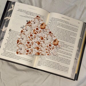 Puede incluir: Libro abierto con un marcapáginas con temática de calabazas. El marcapáginas presenta un patrón de calabazas blancas y naranjas con follaje otoñal. Las páginas del libro son visibles, con texto y un borde decorativo.