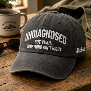 May include: A charcoal gray baseball cap with the embroidered text "UNDIAGNOSED BUT YEAH, SOMETHING AIN'T RIGHT" in white. The cap has a curved brim and a small button on top. The name "Micheal" is embroidered on the side.