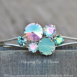 May include: A silver bangle bracelet with a cluster of iridescent pastel green and pink rhinestones. The stones are arranged in a random pattern and are set in silver.
