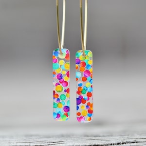 May include: Gold hoop earrings with a colorful confetti design. The confetti is made of small, round, translucent circles in various colors, including red, orange, yellow, green, blue, purple, and pink. The earrings are hanging from a branch.