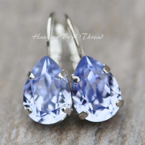 May include: A pair of light blue teardrop earrings with a silver-tone metal setting. The earrings have a faceted, gem-like appearance and are displayed against a blurred background. The text "Hanging By A Thread" is visible.