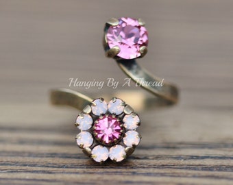 May include: An antique brass-toned ring featuring two pink crystal accents. One is a large, round stone, and the other is a cluster of smaller crystals in a floral design. The ring has a unique, open design with the text "Hanging By A Thread".