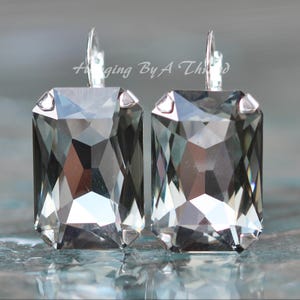 Puede incluir: Un par de pendientes rectangulares de cristal gris con ganchos plateados. Los cristales facetados reflejan la luz. Los pendientes se muestran sobre una superficie reflectante. El texto de la imagen dice "Hanging By A Thread".