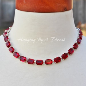 May include: A red rectangular gemstone necklace with silver-colored metal links. The necklace is displayed on a white mannequin. The text "Hanging By A Thread" is visible.