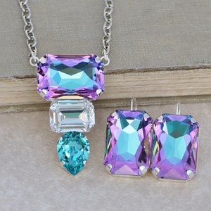May include: A silver necklace and earrings set with iridescent purple, blue, and clear crystals. The necklace features a large rectangular crystal, a smaller rectangular crystal, and a teardrop-shaped crystal.