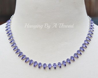 May include: A delicate necklace featuring a series of teardrop-shaped, purple gemstones set in a silver-toned metal. The necklace is displayed on a mannequin, showcasing its design and craftsmanship. The text "Hanging By A Thread" is visible.