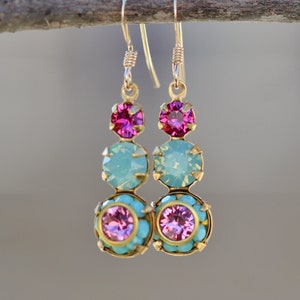 Puede incluir: Pendientes de aro dorados con tres piedras de cristal rosa y azul. Los pendientes están colgados de una rama.
