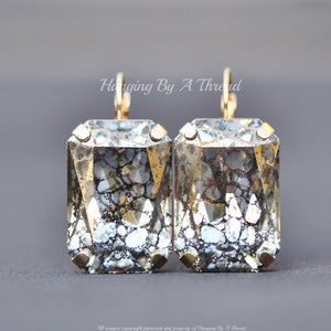 Puede incluir: Un par de pendientes de aro dorados con grandes piedras de cristal rectangulares transparentes. Las piedras tienen un acabado craquelado plateado y dorado moteado.