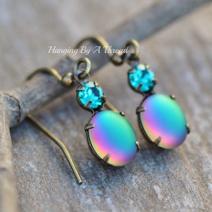 May include: A pair of iridescent green and blue dangle earrings with a small turquoise crystal accent. The earrings have a gold tone finish and are hanging from a hook.