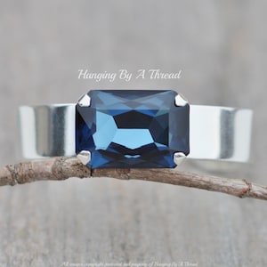 May include: A silver cuff bracelet with a large, dark blue, rectangular gemstone. The bracelet is resting on a branch.