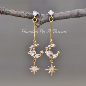 Peut inclure: Boucles d'oreilles pendantes dorées avec un motif de croissant de lune et d'étoile. La lune est ornée de petites pierres scintillantes et l'étoile est une étoile à six branches avec un centre scintillant.