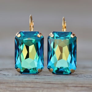 Könnte beinhalten: Ein Paar rechteckige, teal-farbene Ohrringe mit goldfarbenen Metallakzenten. Die Ohrringe haben einen Klappverschluss und ein facettiertes, juwelenartiges Aussehen. Die Ohrringe sind ein Modeaccessoire.
