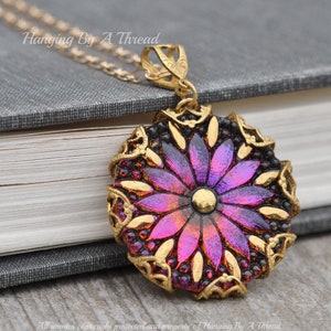 Può includere: Una collana dorata con un ciondolo con un design floreale iridescente nei toni del viola e dell'arancione. Il fiore è racchiuso in una cornice in filigrana nera e dorata. Il testo "Hanging By A Thread" è visibile.