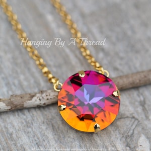 May include: A gold-colored chain necklace with a large, round pendant. The pendant features a vibrant, faceted gemstone with a gradient of colors, transitioning from orange to pink and purple. The text "Hanging By A Thread" is visible.