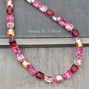 Puede incluir: Un collar de cristal rosa, rojo y dorado con piedras de forma cuadrada. El collar está sobre una superficie gris con el texto "Hanging By A Thread" impreso en él.