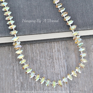 May include: A silver necklace with iridescent, marquise-shaped crystals. The necklace is displayed on a wooden surface with a book in the background. The text "Hanging By A Thread" is visible.