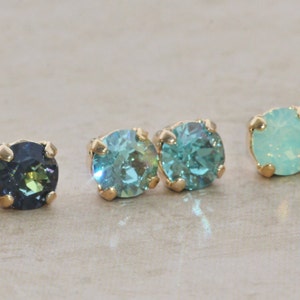 May include: Three pairs of gold stud earrings with different colored gemstones. The first pair has dark blue gemstones, the second pair has light blue gemstones, and the third pair has light blue opalescent gemstones.