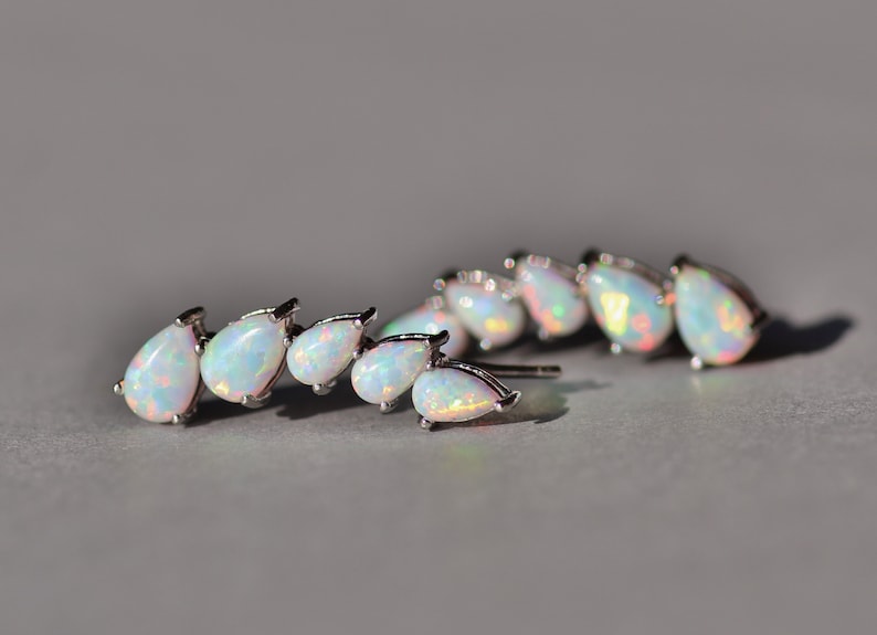 Pu&ograve; includere: Orecchini argentati con un gruppo di opali a forma di goccia. Gli opali hanno una base bianca lattea con lampi di colore iridescente. Gli orecchini sono appoggiati su una superficie grigia.