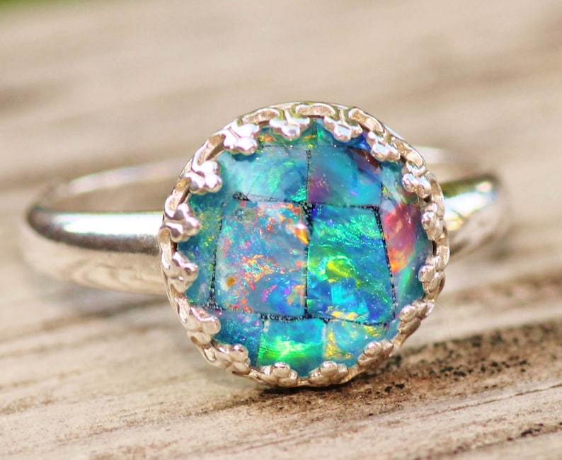 May include: A silver ring with a large, round, mosaic-style gemstone. The gemstone is primarily blue and green with flashes of other colors. The ring's setting has a decorative, scalloped edge.