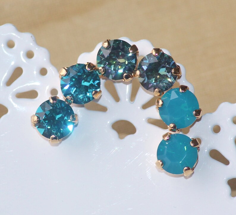 May include: A set of six turquoise and teal blue rhinestones with gold settings. The rhinestones are arranged in a curved shape.