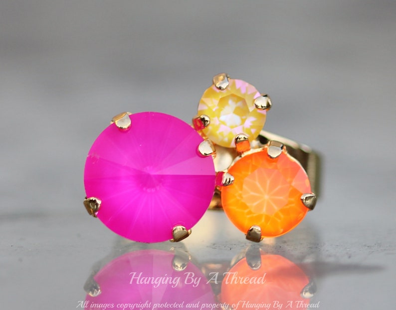 May include: A gold-toned ring featuring three large, round gemstones in vibrant colors: hot pink, orange, and yellow. The gemstones are set with gold prongs. The ring is reflected on a shiny surface. Text: "Hanging By A Thread".