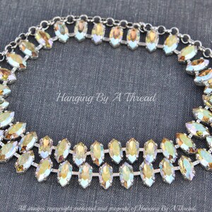 May include: A silver-toned necklace with two rows of iridescent, marquise-shaped crystals. The crystals have a yellow and green color gradient. The necklace is on a dark gray background. The text "Hanging By A Thread" is visible.