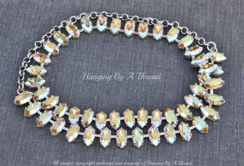 May include: A silver-toned necklace with two rows of iridescent, marquise-shaped crystals. The crystals have a yellow and green color gradient. The necklace is on a dark gray background. The text "Hanging By A Thread" is visible.